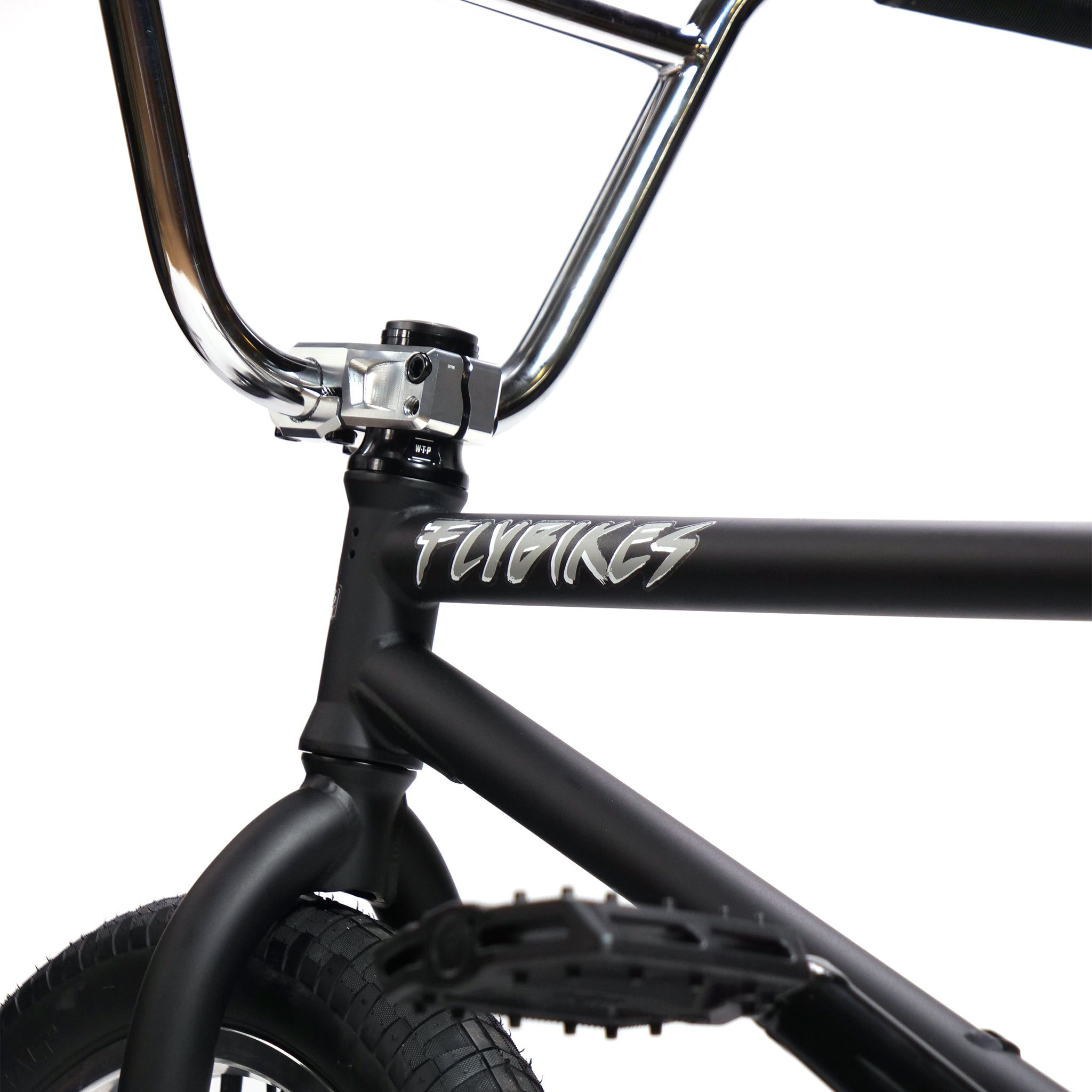 Close-up of the Fly Bikes Pajarito Custom 18 Inch BMX Bike, showcasing a black frame with "FLYBIKES" in white. Visible parts include the handlebar, frame junction, front wheel, and pedal.