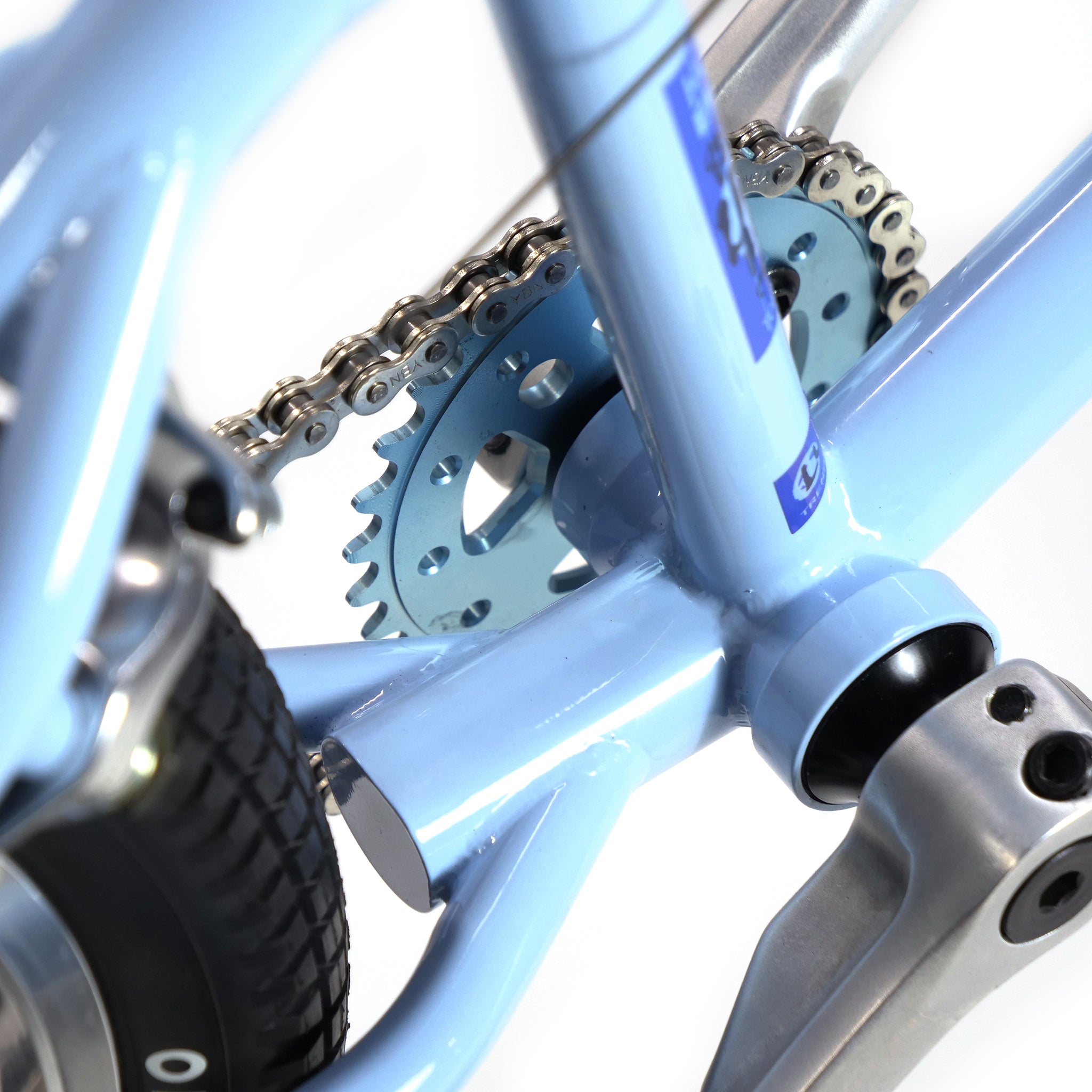 Close-up view of the chain and sprocket on a light blue Hoffman TAJ Trend Adjacent Joy 20 Inch Custom Bike, featuring BMX freestyle flair, with part of the rear tire and pedal visible.