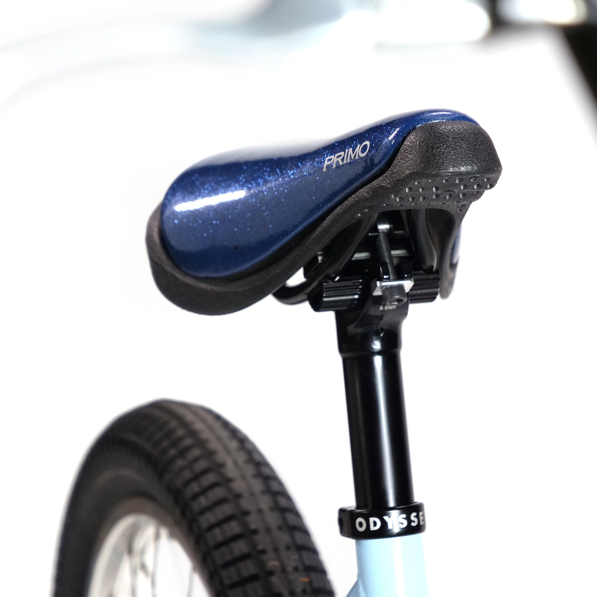 Close-up of a blue "PRIMO" seat on a black support post, showcasing Hoffman Bikes' BMX freestyle touch, with the rear wheel featuring intricate tire tread. Product: Hoffman TAJ Trend Adjacent Joy 20 Inch Custom Bike.