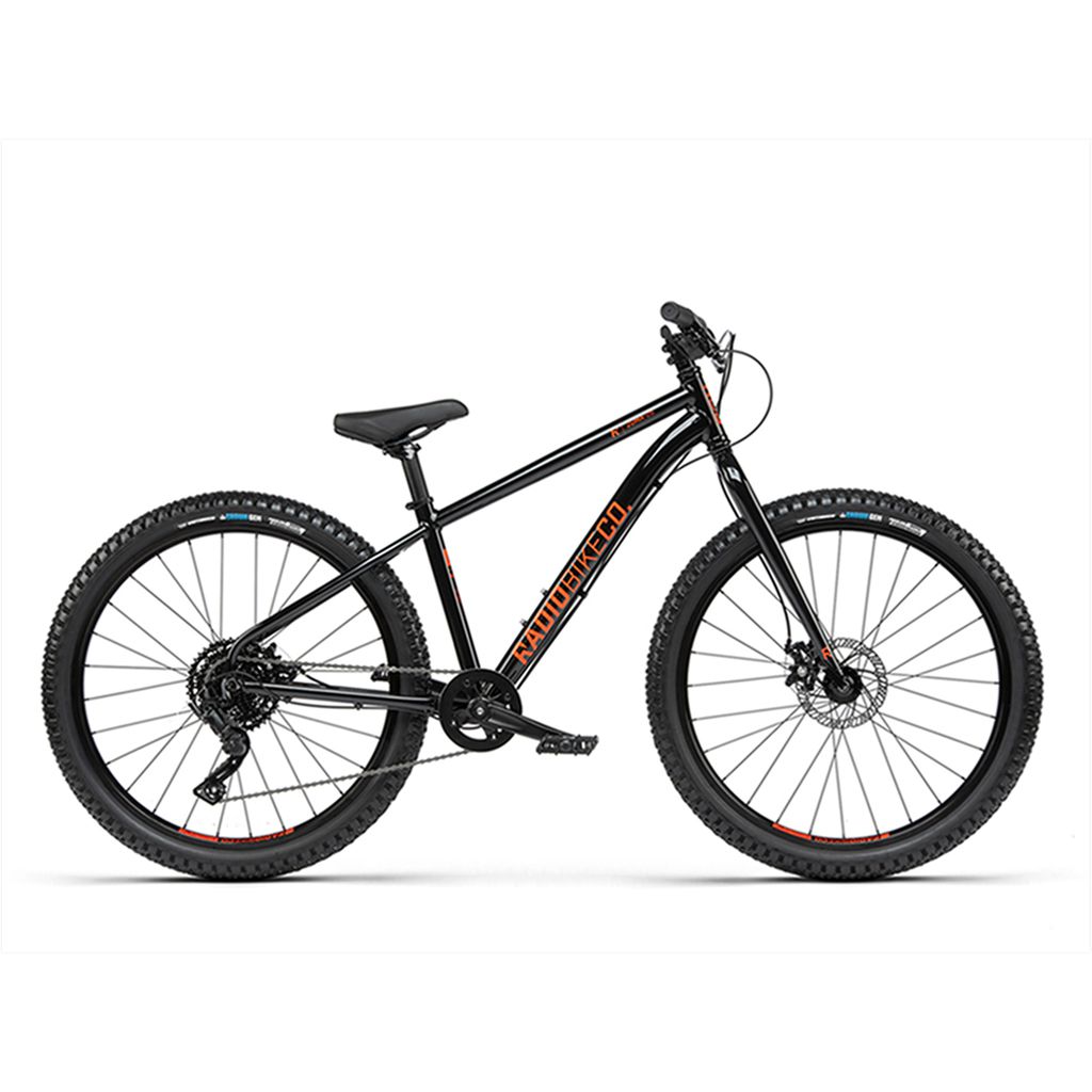 The Radio 26 Inch Zuma Bike is a black kids' MTB featuring a lightweight alloy frame, large knobby tires, front suspension, disc brakes, and an orange brand logo on the frame.