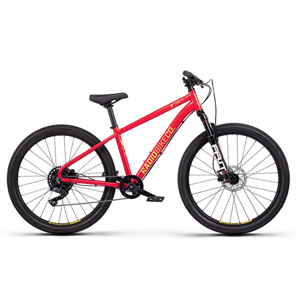 The Radio 26 Inch Zuma Sus Bike is a red kid-sized MTB bike featuring a sturdy frame, front suspension fork, and knobby tires. It includes a black saddle, pedals, handlebars, and a gear system built on a lightweight alloy frame.