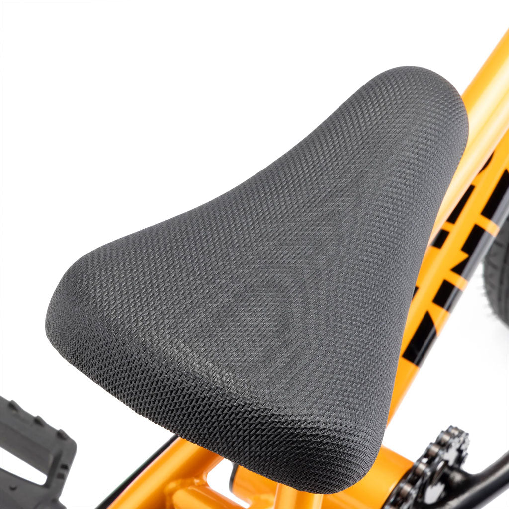 Close-up of a black textured bicycle seat mounted on an orange bike frame, with part of a pedal visible beside it—perfect details for a Kink Roaster 12 inch Bike (2025).