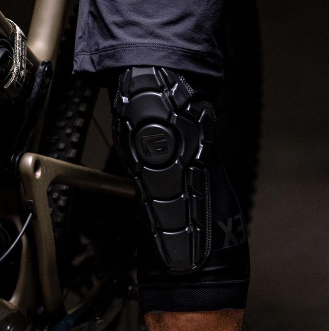 A close-up shows a person wearing G-Form Pro-X3 Knee Guards with SmartFlex protection while sitting on a bike, highlighting their preparedness for impact-absorbing adventures. The bike frame and part of the tire are also visible.