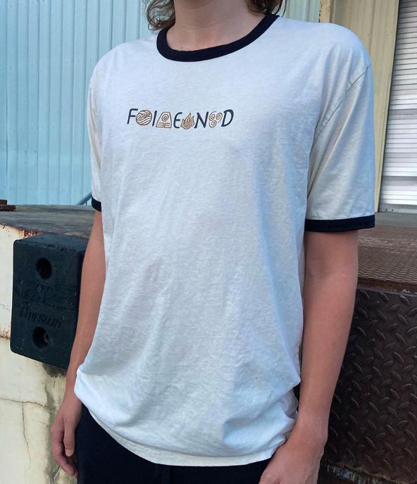 A person wears the Fiend Shirt Raekes DT Natural— a ringer-style t-shirt with a white body and black trim, featuring abstract text. The setting is an industrial backdrop with a metal wall, adding an edgy vibe typical of rider-owned brands.