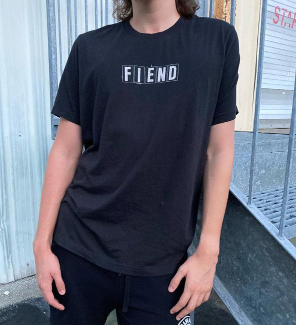 A person in a black Fiend Ransom Vintage Shirt, made from 100% airlume cotton with "FIEND" stylishly discharged printed on it, poses confidently before a metal fence.