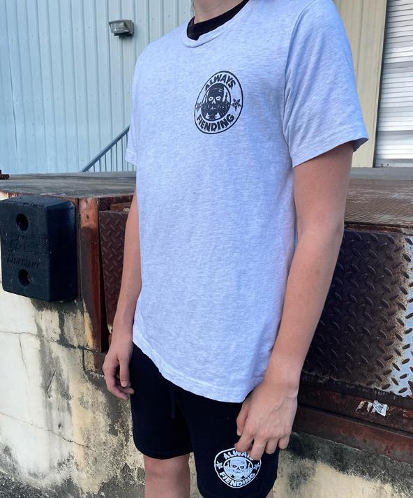 A person wearing a Fiend Always Fiending Shirt (Ash) and black shorts stands near a building with a metal dock plate outdoors.