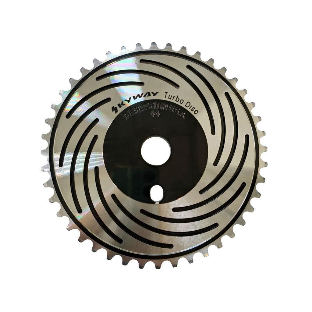 A shiny metallic disc, CNC machined from 6061 Aluminum, with gear-like teeth around its edge, labeled "Skyway Turbo Disc Front Sprocket." The disc features a swirl pattern of slotted holes and two central holes.