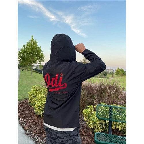 In a park surrounded by trees, grass, and a bench, someone wears an ODI Heater Windbreaker with red text on the back of their black hoodie. Their water-resistant jacket fits perfectly with the peaceful setting as they stand turned away.