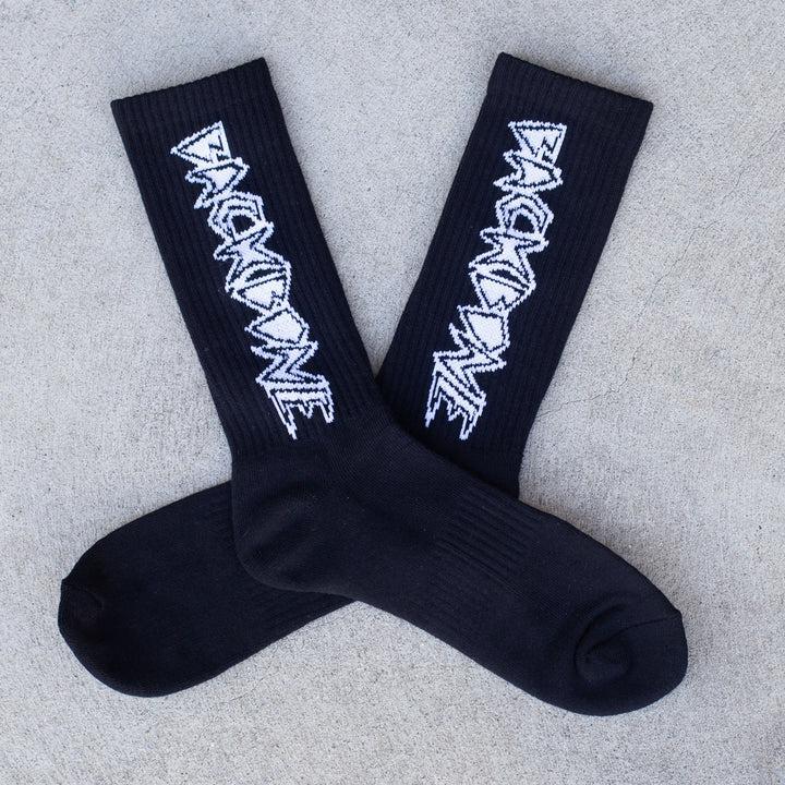 The Backbone Crew Socks are buttery soft, black with a white zigzag text design, and stylishly placed crosswise on a concrete surface. These one-size-fits-most socks combine comfort and flair.