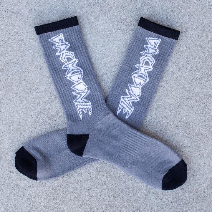 Backbone Crew Socks in gray, crafted with buttery soft fabric, feature black toe and heel areas along with a white stylized logo on the sides. They are placed on a concrete surface and come in one size that fits most.