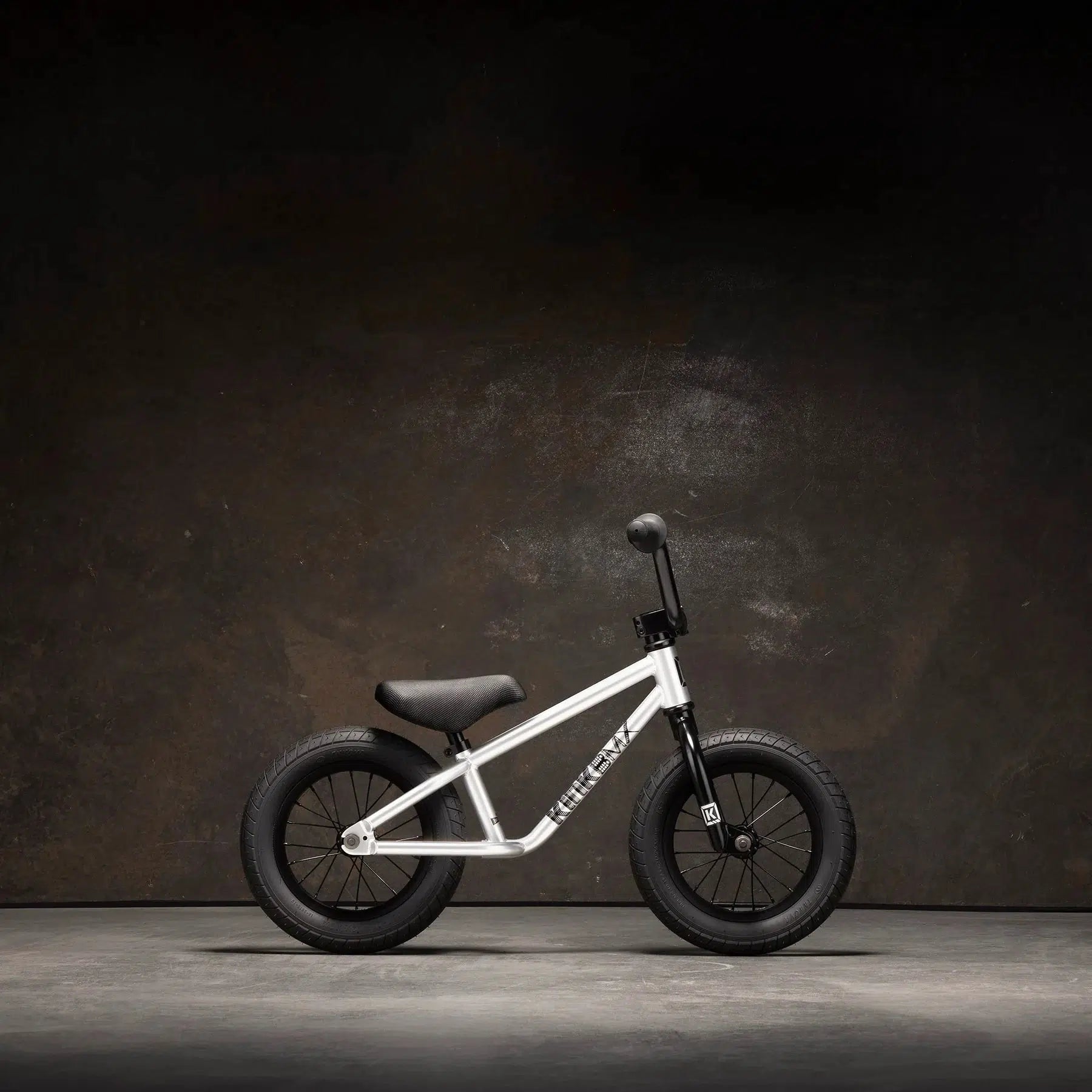 The Kink Coast Balance Bike showcases a silver frame complemented by black wheels, seat, and handlebars, all beautifully highlighted against a dark backdrop.