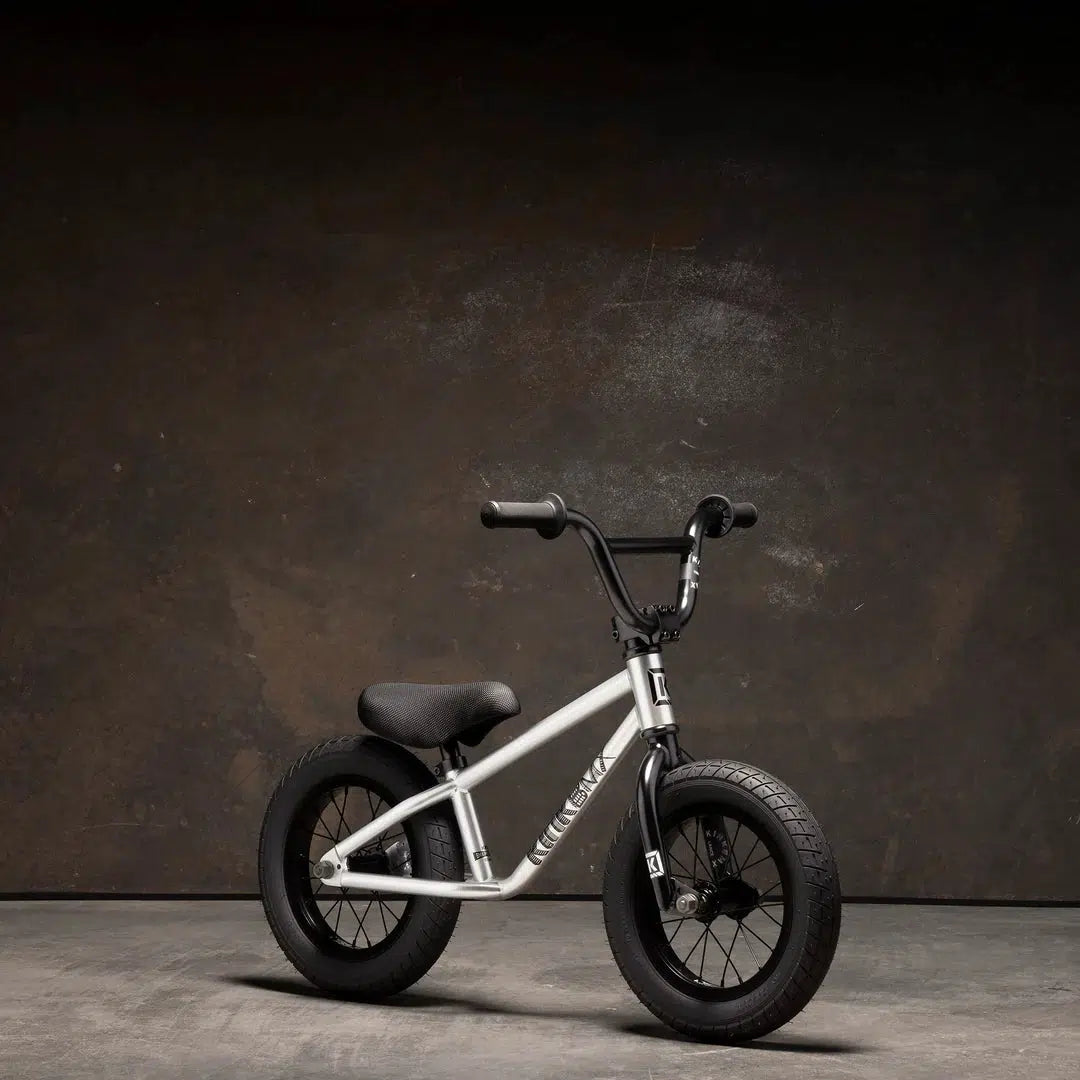 A compact Kink Coast 12 Inch balance bike, featuring a black and silver finish and thick tires, stands out against a dark background.