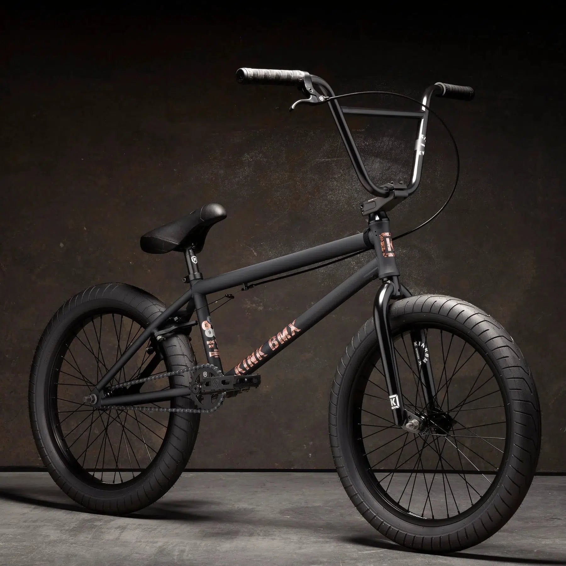 A Kink Gap XL 20 Inch Bike, boasting a sleek black design with modern geometry, thick tires, and a curved handlebar, stands proudly on a matte surface against a dark background. Its 4130 Chromoly frame ensures both strength and style for every ride.