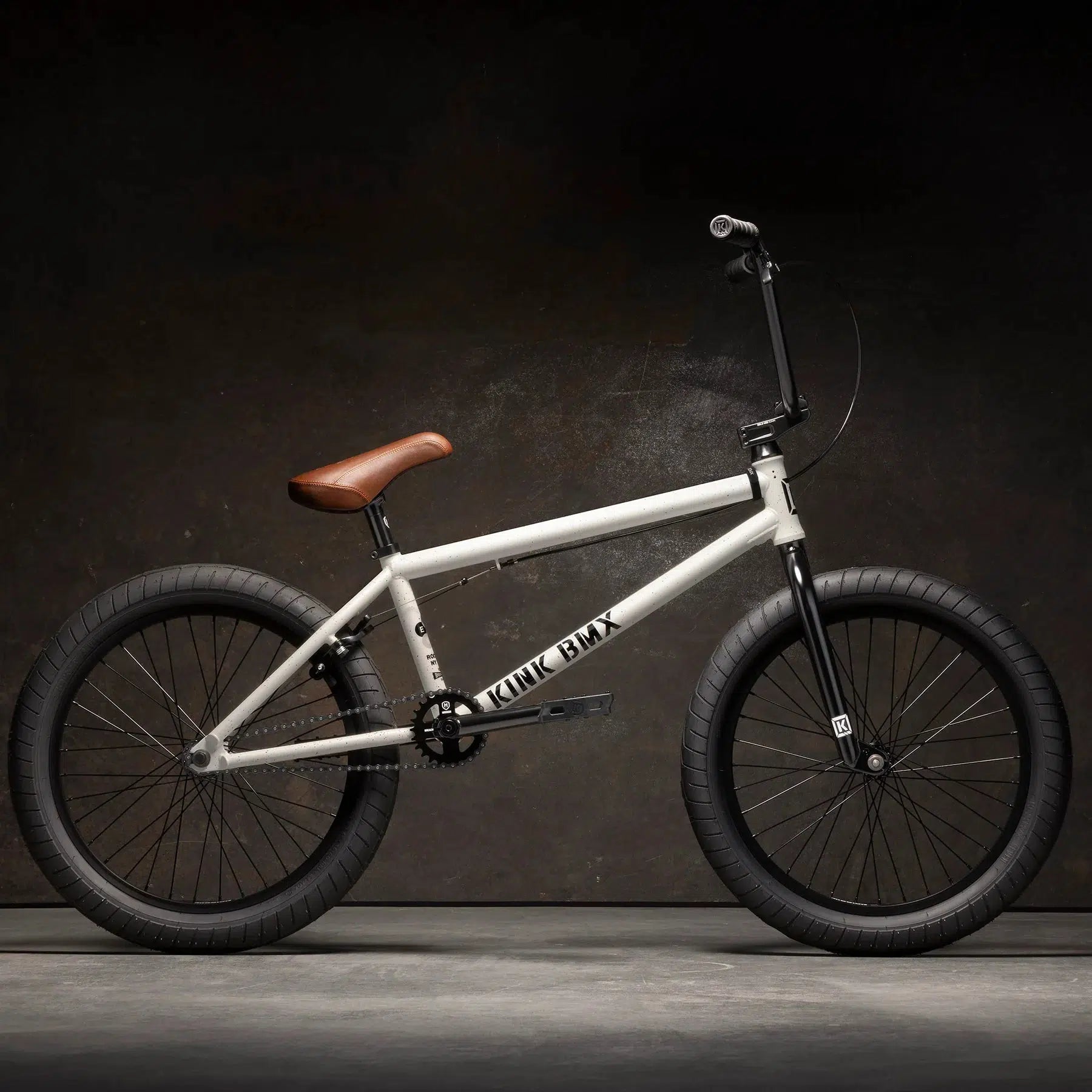 A Kink Gap XL 20 Inch Bike, featuring a white frame, brown seat, and black handlebars and tires, stands boldly against a dark background. Its sturdy 4130 Chromoly construction ensures durability for every adventure.