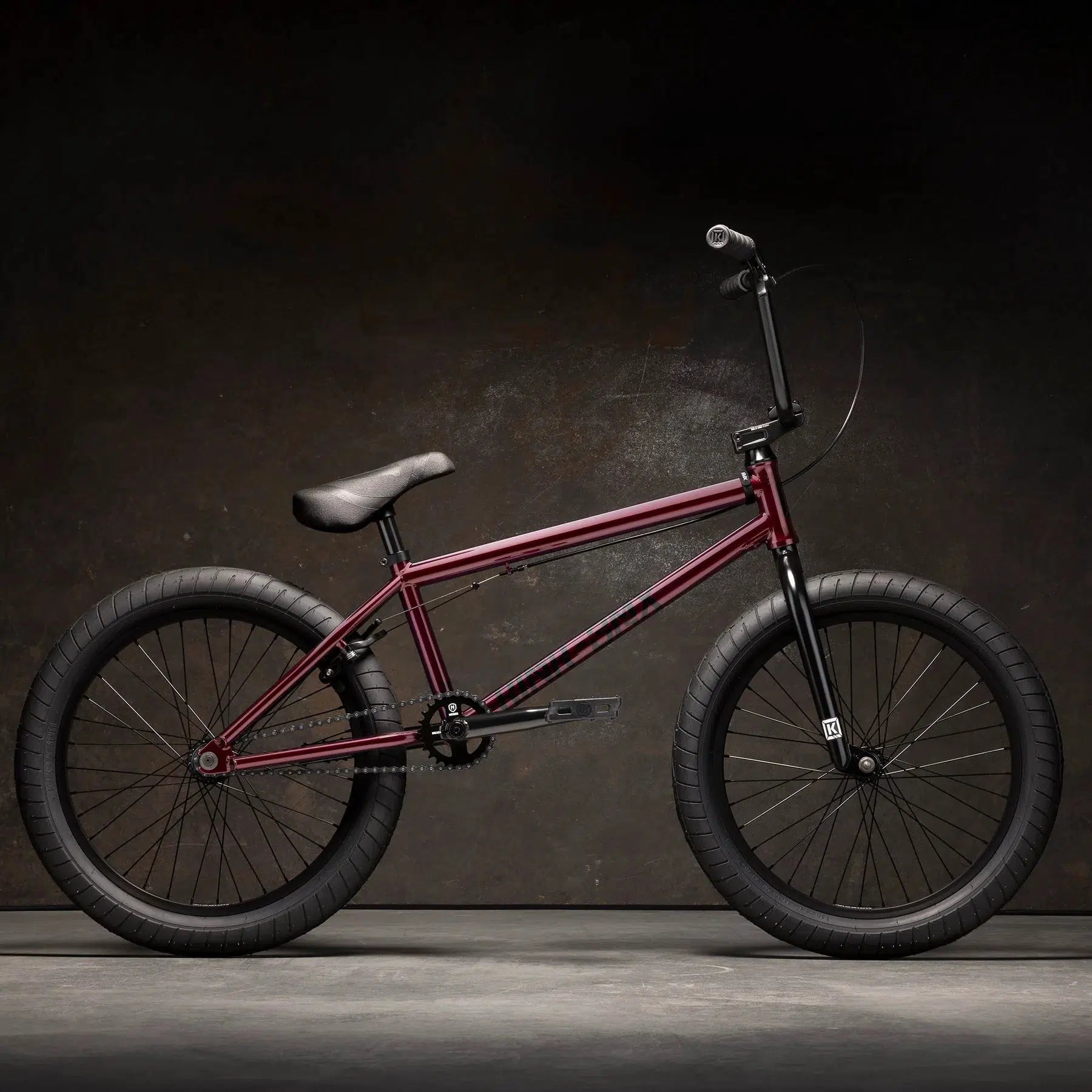 A maroon Kink Launch 20 Inch Bike featuring black handlebars and wheels is displayed against a dark background.