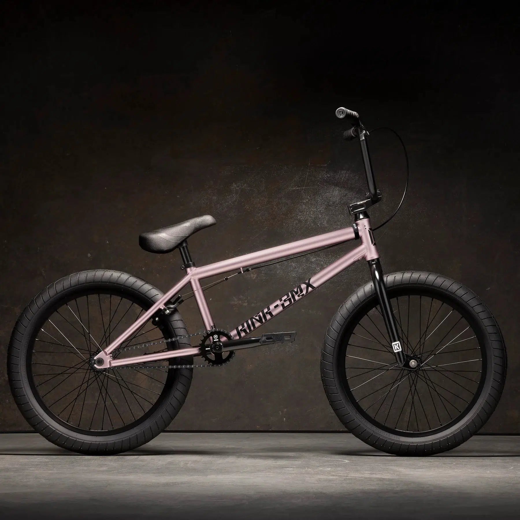 The 2023 Kink Launch 20 Inch Bike boasts a pink frame, thick black tires, and a sleek black seat set against a dark background, enhanced by a sealed integrated headset for smooth steering.