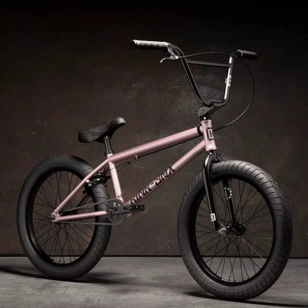 A Kink Launch 20 Inch Bike in pink, featuring black tires and handlebars, is showcased against a dark background.