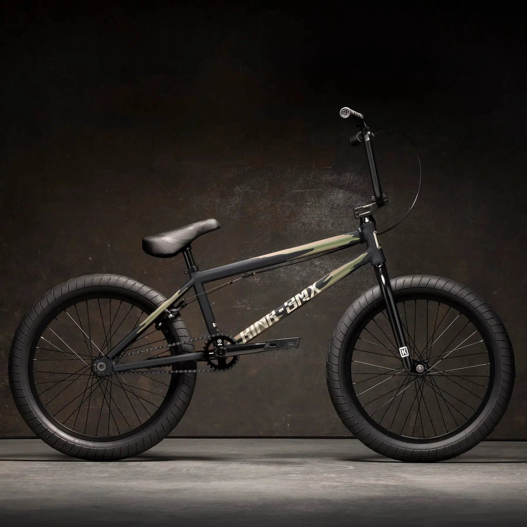 The Kink Launch 20 Inch Bike, a sleek black BMX featuring thick tires and a camouflage-patterned frame, is showcased against a dark background.