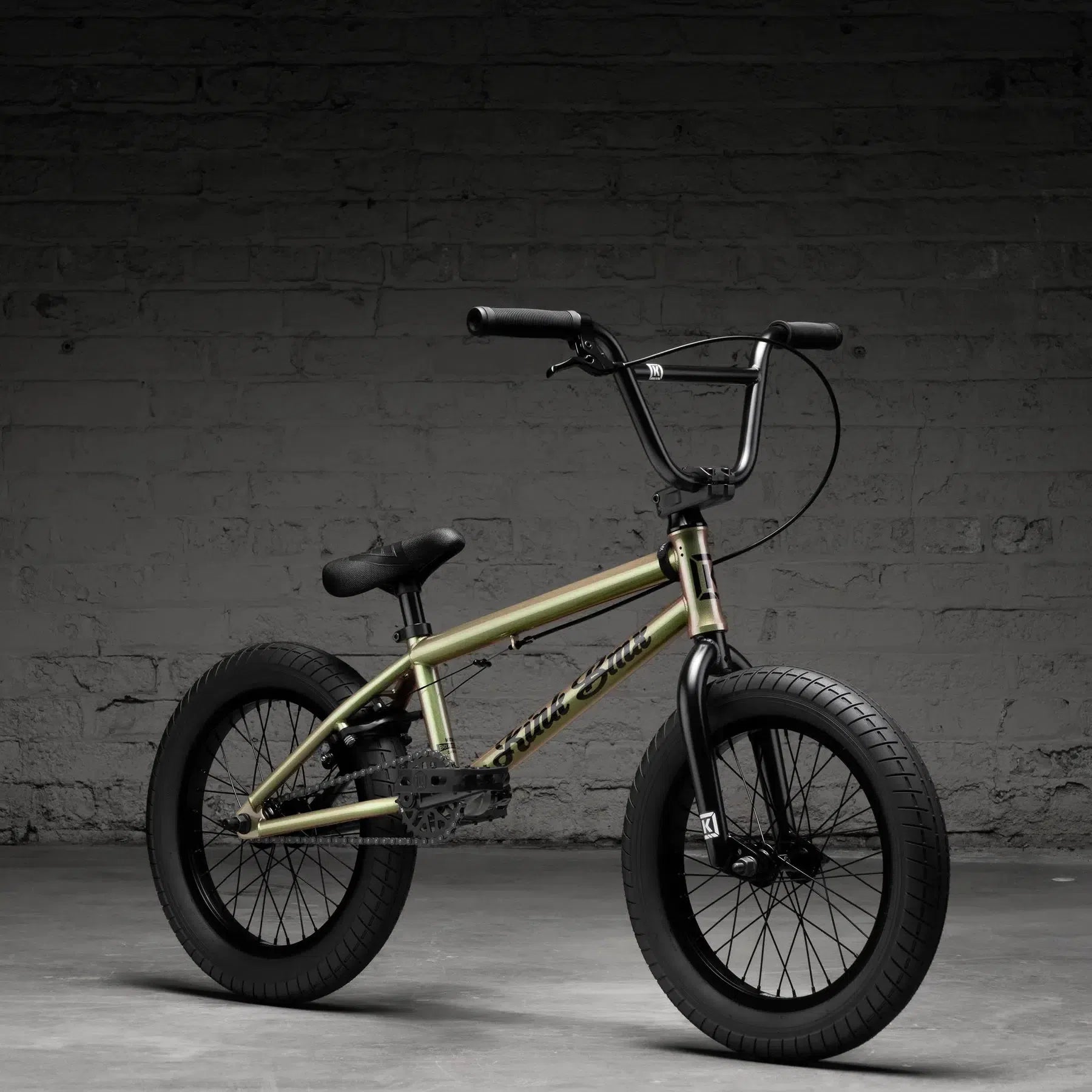 The Kink Carve 16 Inch Bike (2026), featuring a gold frame and black wheels, boldly stands against a dark, textured brick wall, embodying its daring spirit.