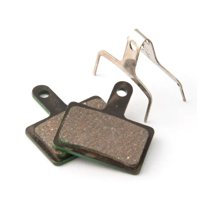 The Clarks VX Organic Disc Brake features a pair of rectangular brake pads with attachment holes, complemented by a metal spring clip in the background, offering improved braking performance and noise reduction for a smoother, quieter cycling experience.