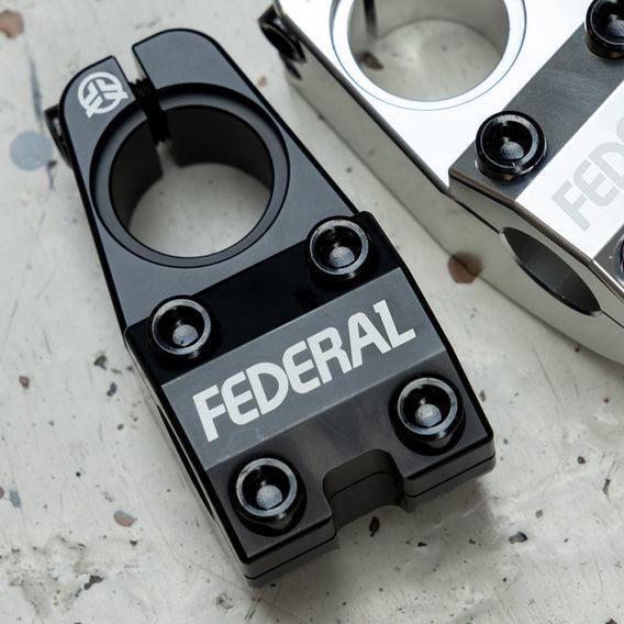 Close-up of two BMX bike stems on a speckled surface; one black with "Federal" text is the Federal Session Topload Stem, and the other is silver, crafted from 6061-T6 Aluminum. The silver stem features CNC precision for optimal weight reduction.