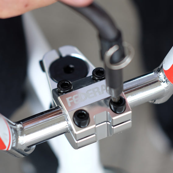 A hand adjusts a Federal Session Topload Stem on a silver bicycle handlebar. Made from 6061-T6 Aluminium, it boasts black bolts and "Federal" engraving, offering durability and reduced weight for top performance.