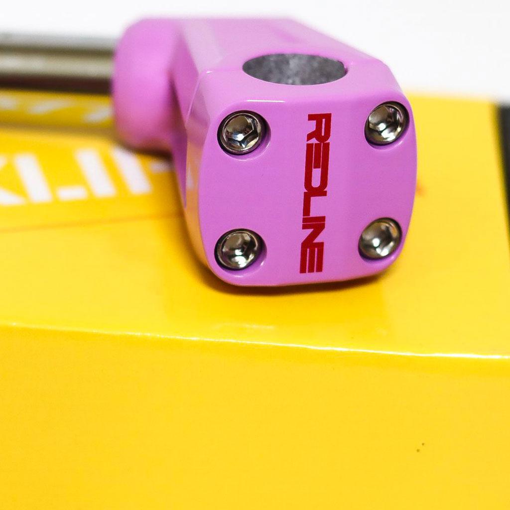 Close-up of a pink scooter clamp with "REDLINE" written in red text, resting on a yellow surface. Four bolts are visible on the clamp, reminiscent of the precision found in BMX Freestyle gear like the REDLINE BY KASTAN Forklifter Front Load Stem.