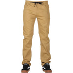 A person stands wearing FP Pants Relaxed Fit Chino Tan featuring a black drawstring. Complementing the outfit, they sport black and white skate shoes with brown soles.