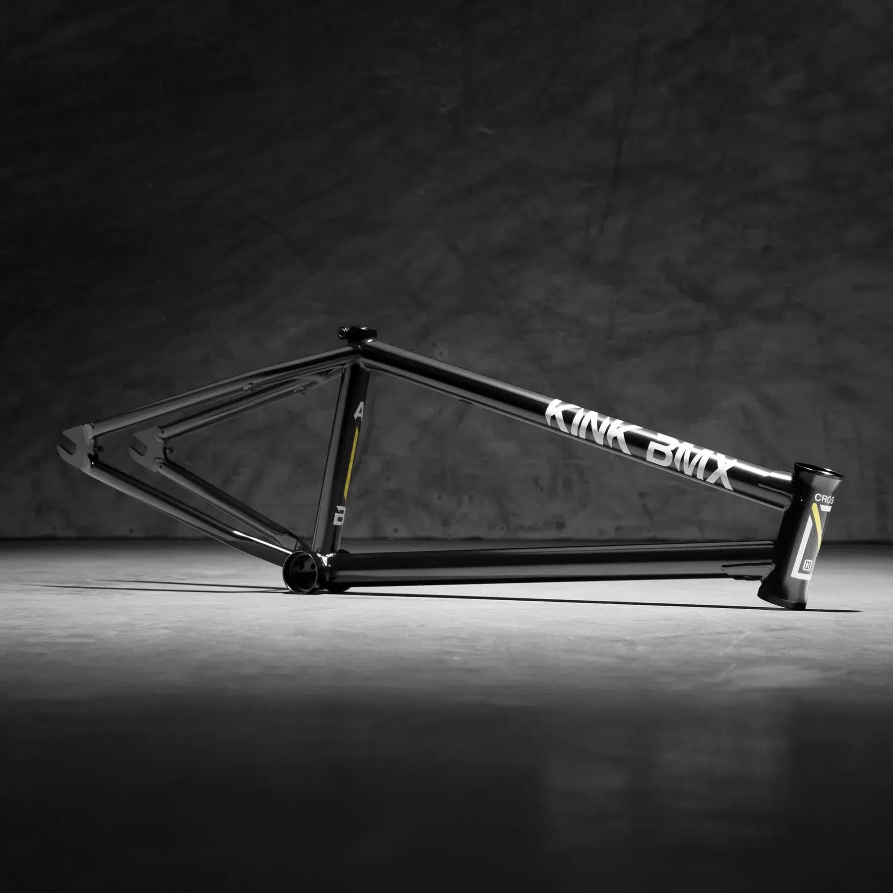 The Kink Crosscut Frame, a sleek black bicycle frame with white branding, rests on a concrete surface. Its modern-day geometry is highlighted against the dark textured background, and it features integrated chain tensioners for optimal performance.