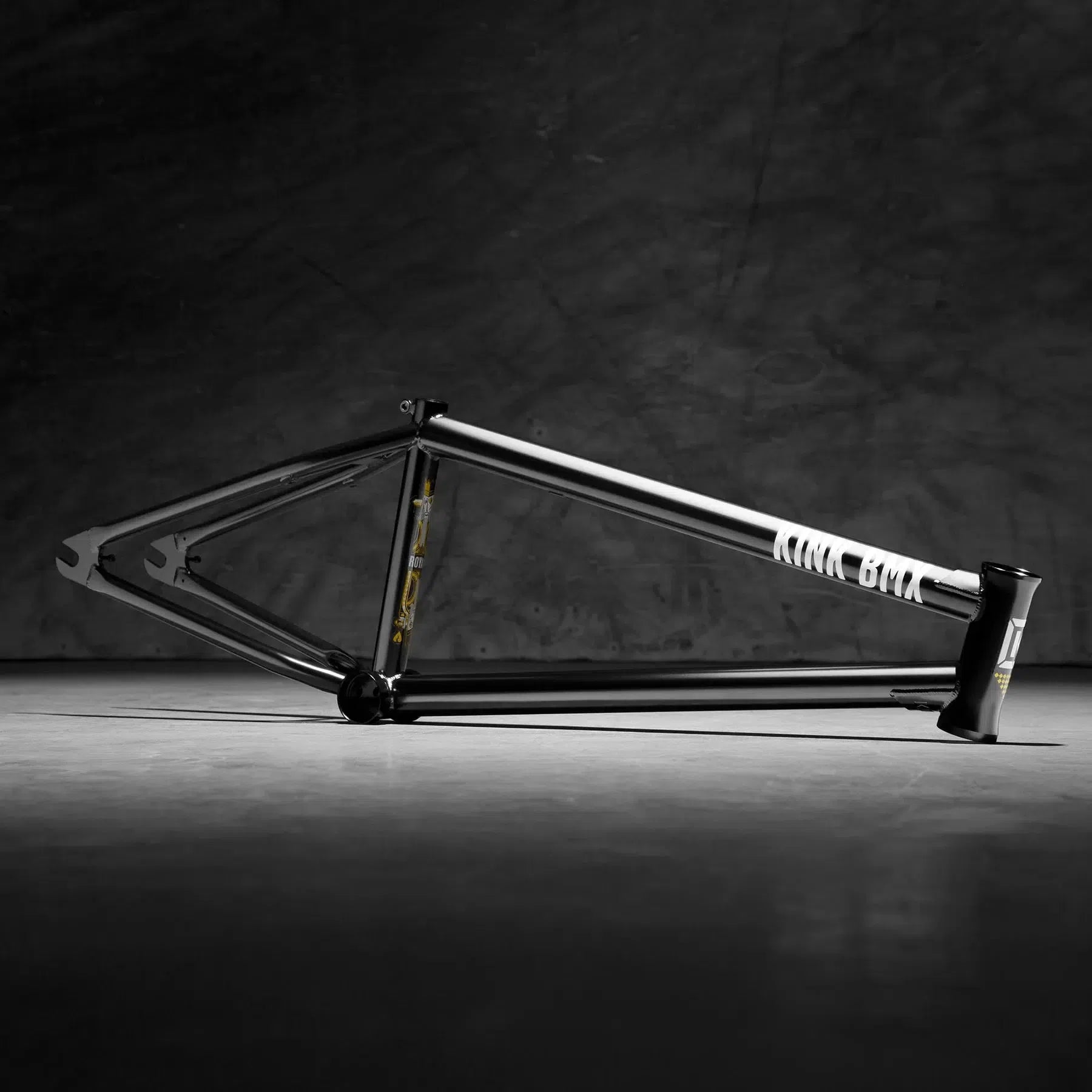 The Kink Royale Frame, made from sturdy 4130 chromoly tubing, rests on a concrete floor against a dark backdrop. Its black and white design features integrated chain tensioners, enhancing both its style and functionality.