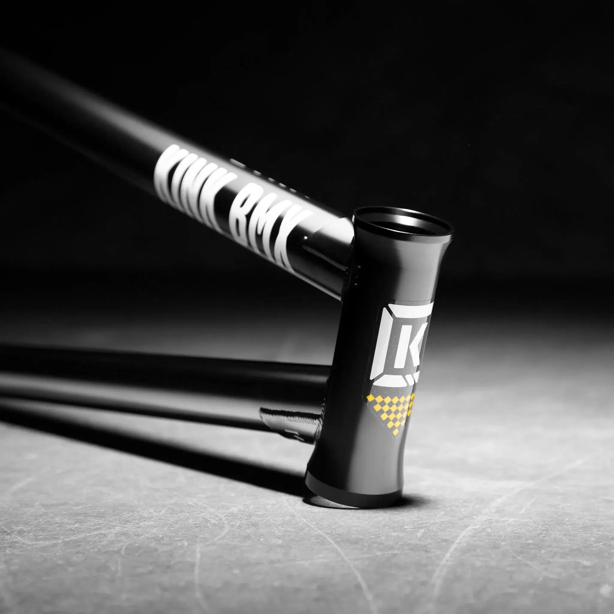 Close-up of a Kink Royale Frame BMX bike in black, featuring branding logos and made from durable chromoly tubing, positioned diagonally on a lit concrete surface—ideal for street and plaza riding.