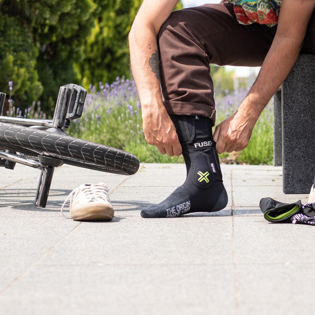 A person is seated on a bench, putting on a black sock with "FUSE" and "THE ORIGIN" printed on it. Nearby, a bicycle and a shoe lie on the ground, seemingly discarded in favor of adjusting the Fuse Omega Pro Ankle Guard.