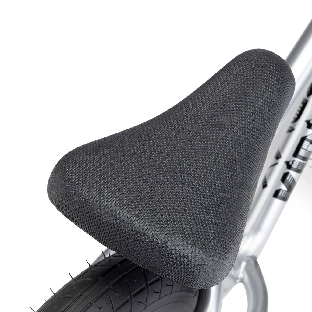 This is a close-up of the black textured seat attached to the silver frame of the Kink Coast Balance Bike, showcasing its sleek BMX-inspired design, with a portion of the rear tire visible.