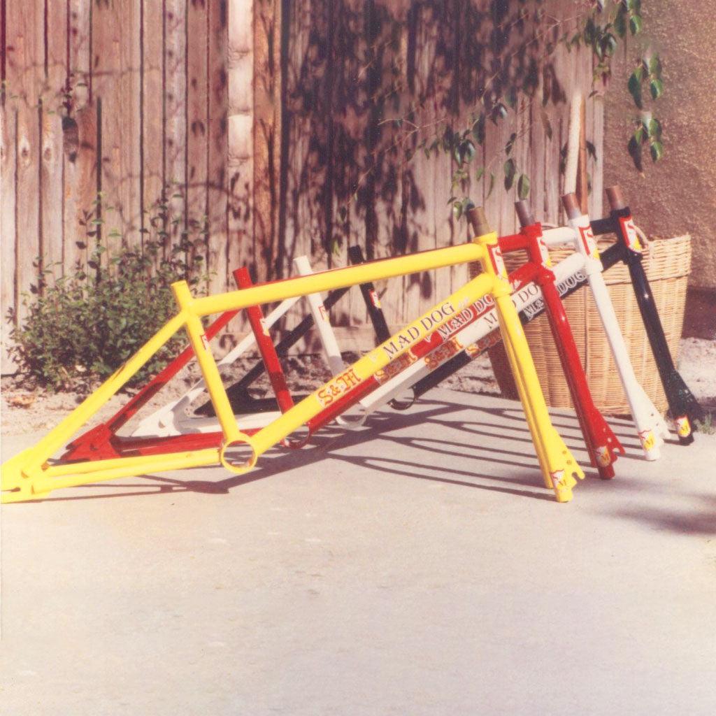 The S&M Mad Dog (Chris Moeller) Disc 20 Inch Frames, featuring sleek designs and disc brakes, are lined up against a wooden fence on a sunny day, ready for any adventure.