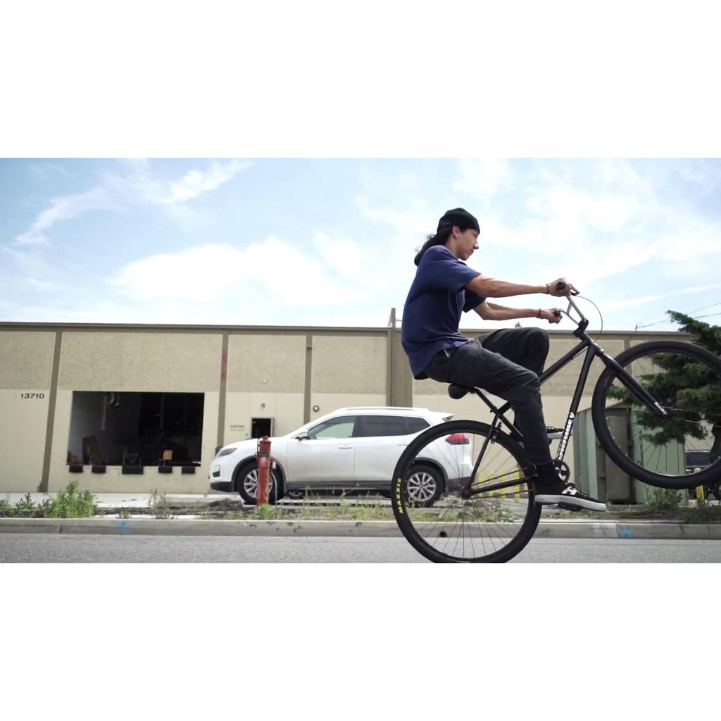 A person skillfully performs a wheelie on a sleek Sunday High C 29 Inch Bike, gliding past a building and parked car in the background.