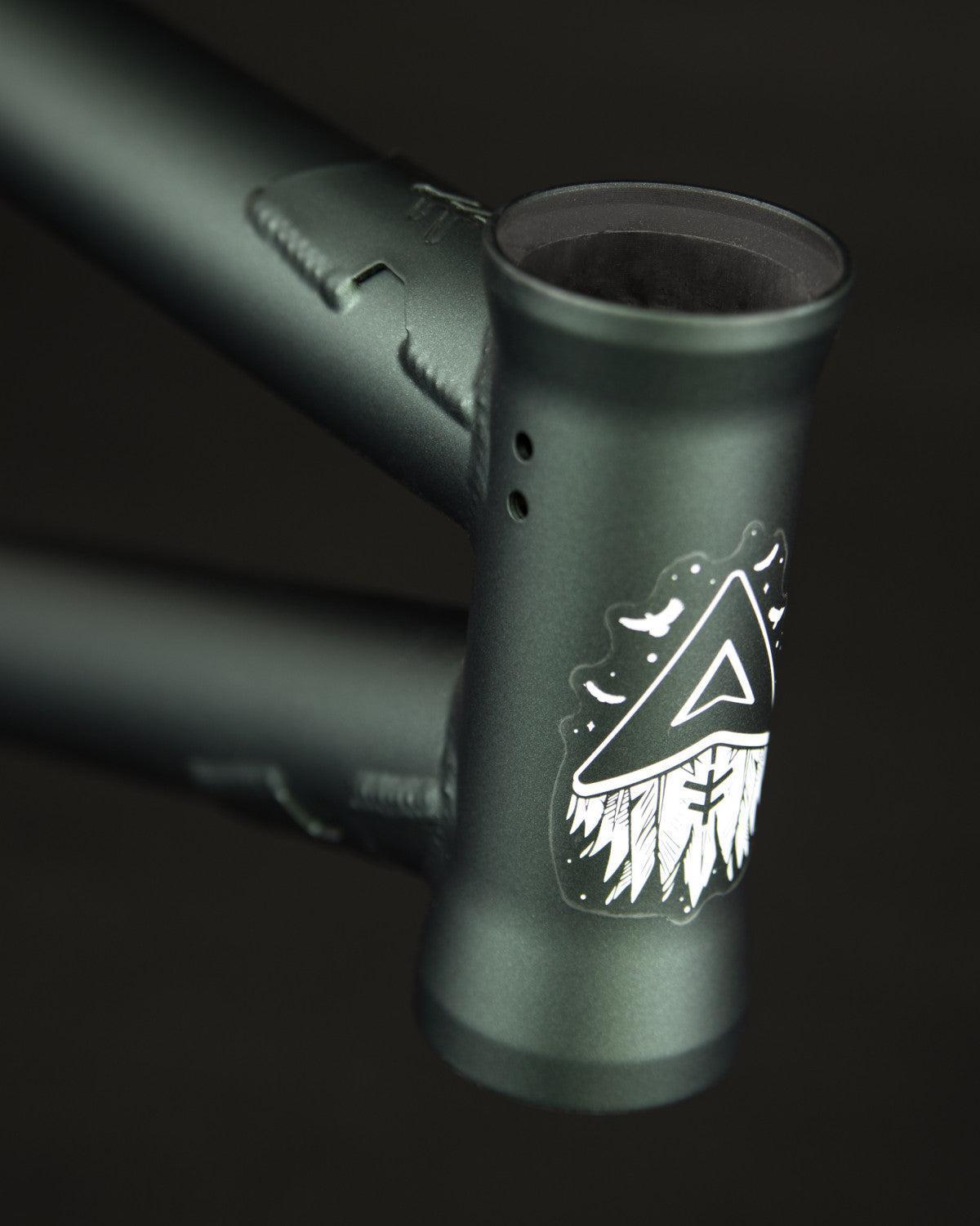 Close-up of the 4130 Cr-Mo Fly Bikes Aire Frame head tube in matte green, displaying a white triangle and stylized feathers graphic on the front.