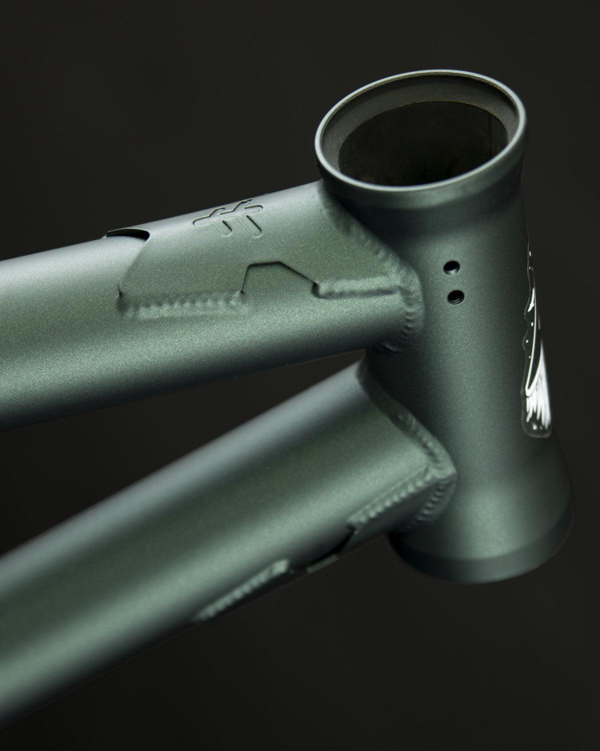 Close-up of a matte green Fly Bikes Aire Frame, highlighting the head tube and welds against a dark background.