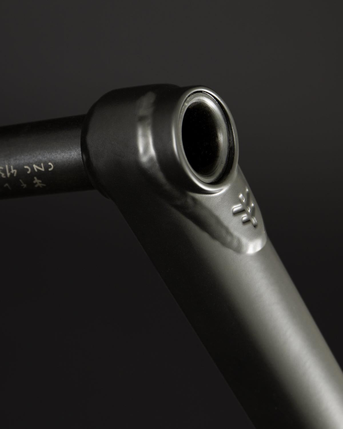 A close-up showcases the Fly Bikes Dolmen 24 Cranks, a sleek black metal part, likely CNC machined from 4130 Chromoly. It features a cylindrical opening and detailed engraved markings set against a dark background.