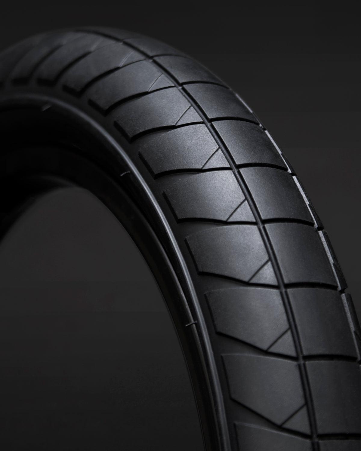 Close-up of the Fly Bikes Fuego Tyre, a black street tire with a geometric tread pattern, showcased on a dark background.