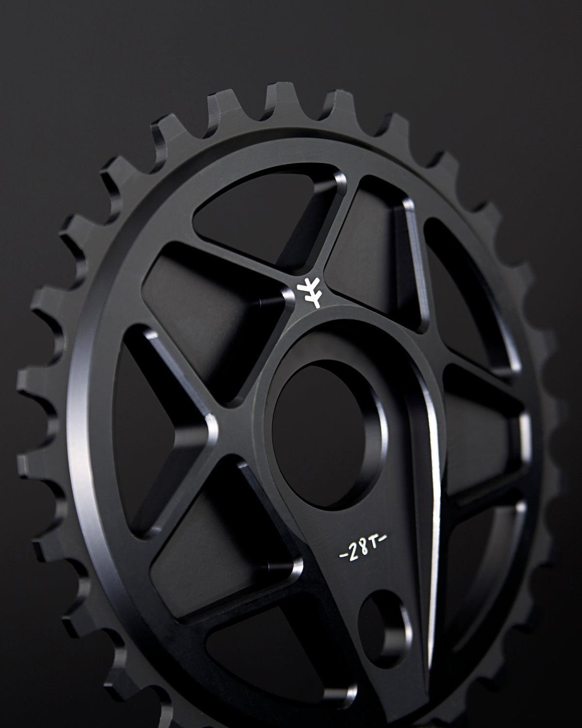 Close-up of the Fly Bikes Tractor XL Sprocket, a black bicycle chainring with 28 teeth, CNC machined from durable 6061 Aluminum, featuring geometric cutouts against a dark background.