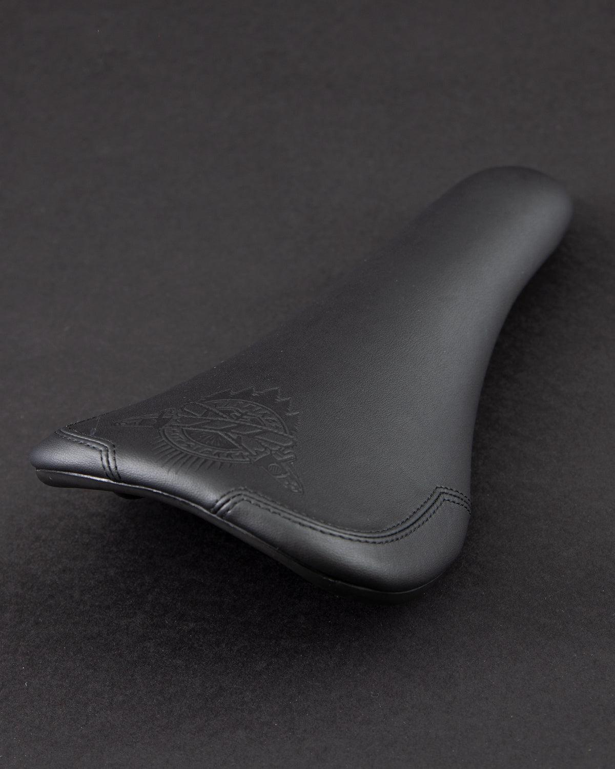 The Motosierra Railed Seat by Fly Bikes is a black seat with a textured logo on a dark background, made from synthetic leather and designed with a railed base for added stability.