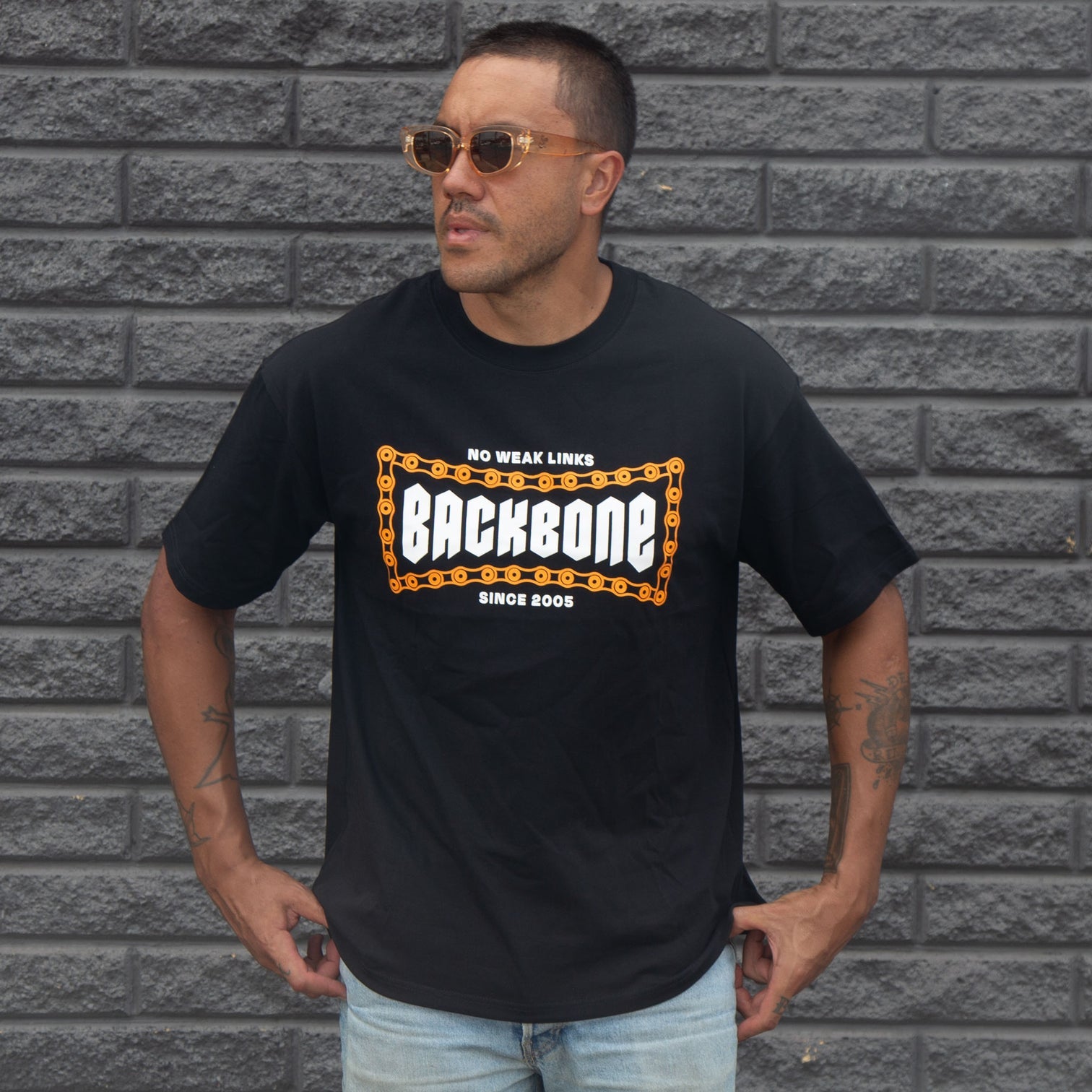 A man wearing sunglasses and a black "Backbone No Weak Links" T-shirt with an orange chain graphic stands against a gray brick wall, highlighting the shirt's boxy fit and heavyweight cotton.