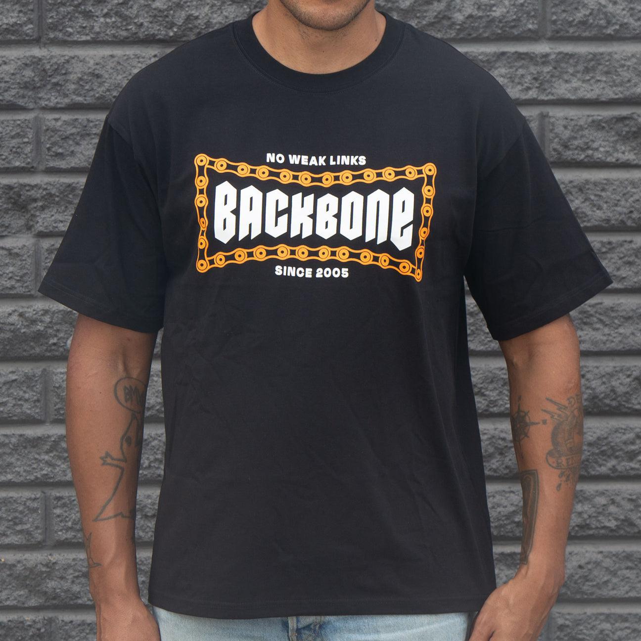 A person wearing the Backbone No Weak Links T-Shirt, styled in a boxy fit. This black heavyweight cotton shirt features "BACKBONE" encircled by a chain design, with "No Weak Links" above it, set against a brick wall backdrop.