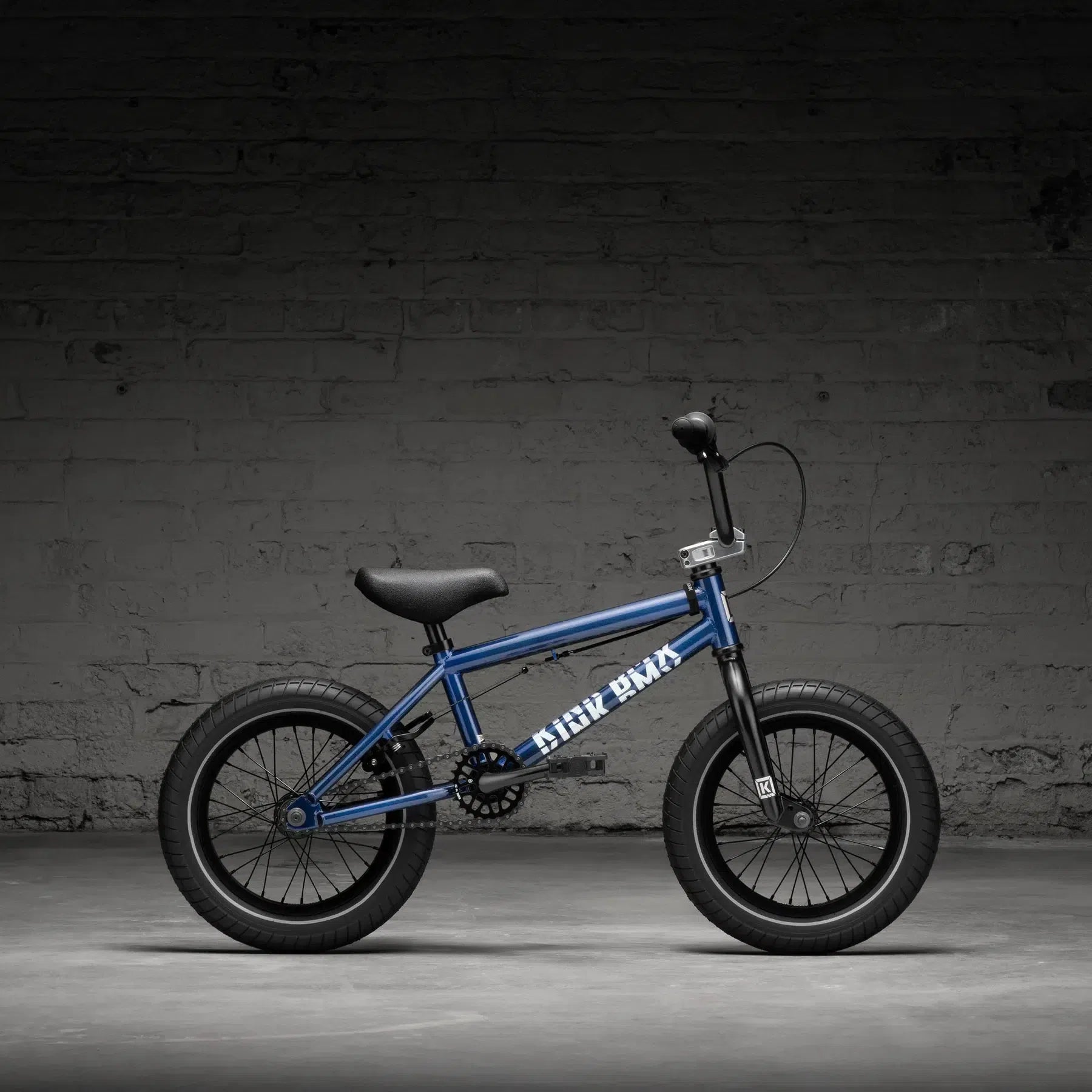 A Kink Pump 14 Inch Bike (2026) in blue, featuring a black seat and tires, is displayed against a gray brick wall.