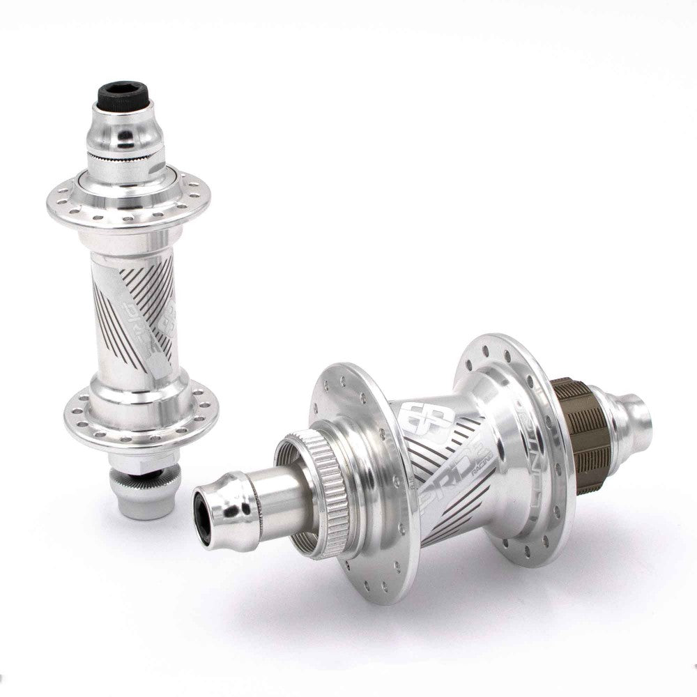 The Pride Control Expert 28H Hubset consists of two expertly crafted silver hubs made from CNC machined aluminium, featuring intricate engravings. One hub stands upright while the other lies on its side, demonstrating their BMX racing readiness with disc brake compatibility. They are elegantly displayed on a white background.