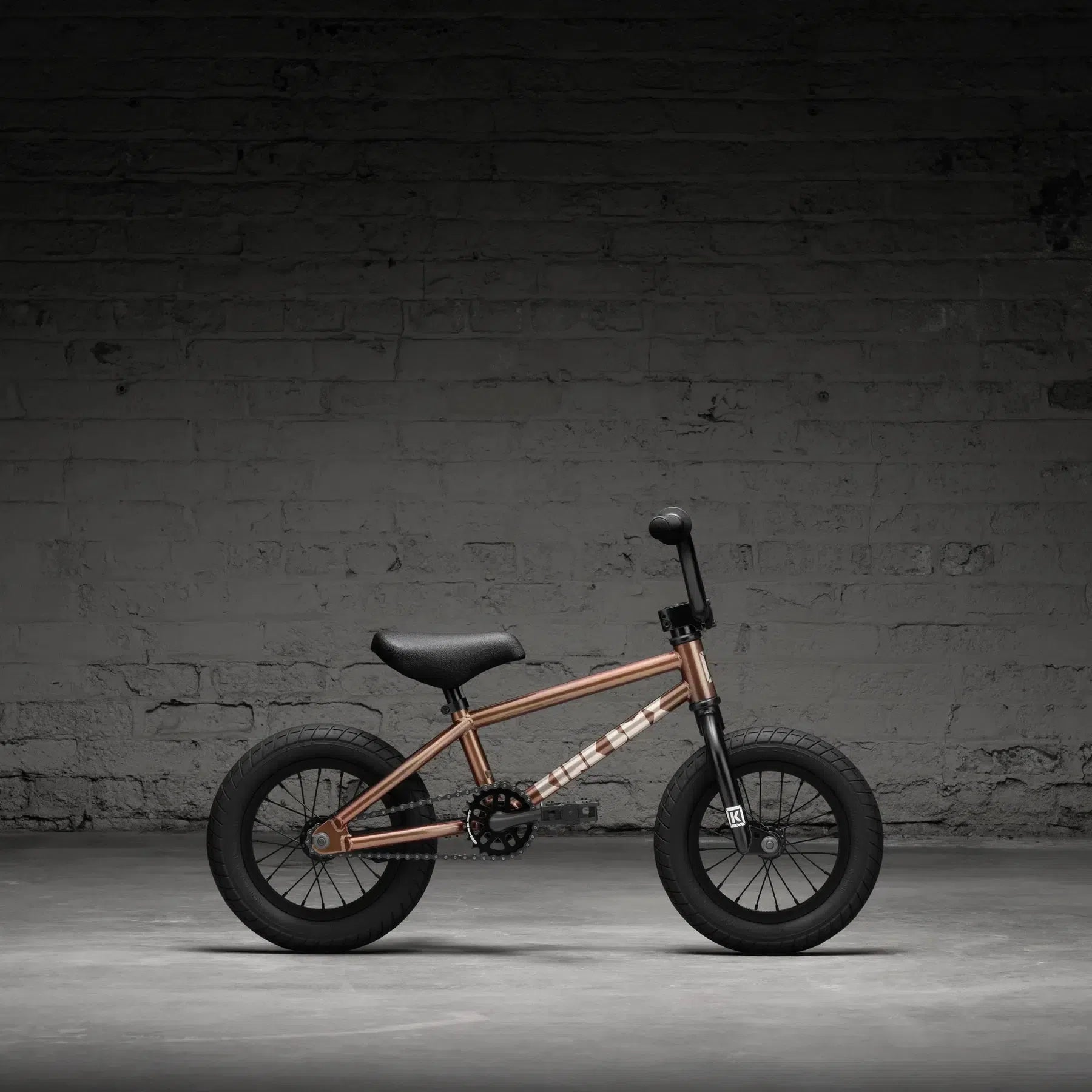 The Kink Roaster 12 Inch Bike (2026), featuring a bronze frame and black wheels, is ideally suited as a child's first BMX and is set against a dark brick wall.