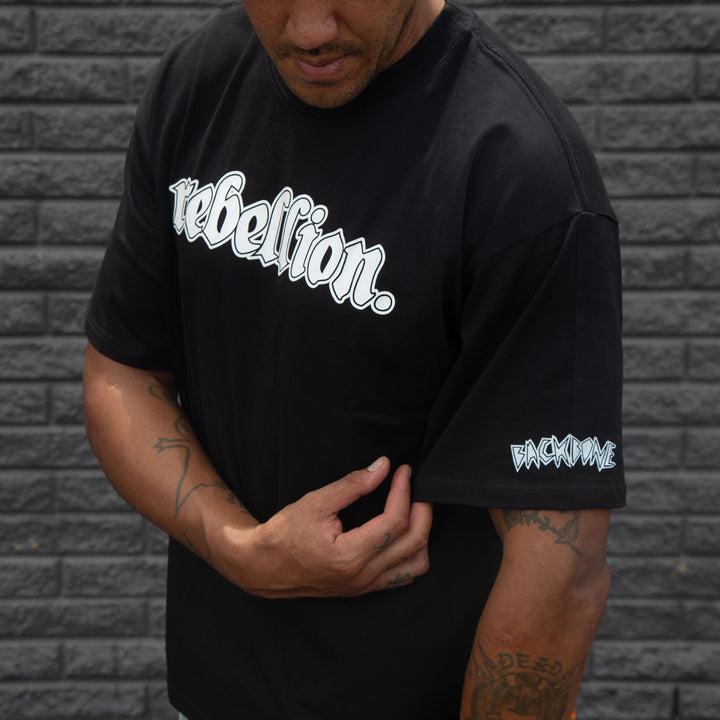 Against a gray brick wall, a person wears the black Backbone Rebellion T-Shirt, boxy fit and crafted from pre-shrunk cotton, featuring "rebellion" in bold on the front and "backbone" on the sleeve.